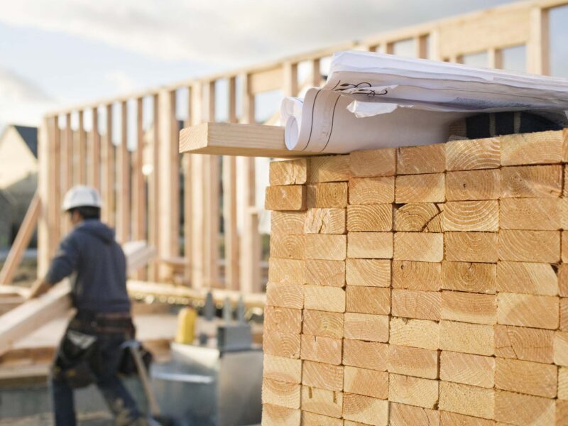 A closeup of stacks of 2x4 boards at a construction site, with a roll of blueprints sitting on top. Two construction workers and a building frame can be seen in the background. Horizontal shot.
